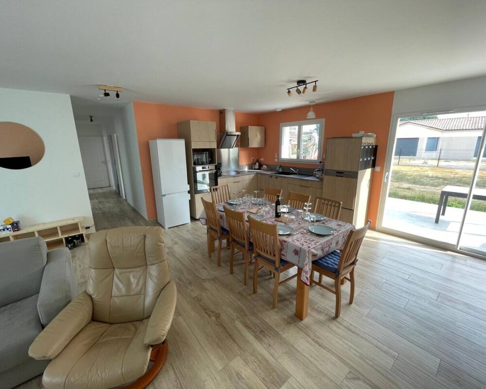 Cuisine ouverte moderne avec meubles bois et murs orange. Grande table a manger, refrigerateur, fauteuil en cuir et acces terrasse.