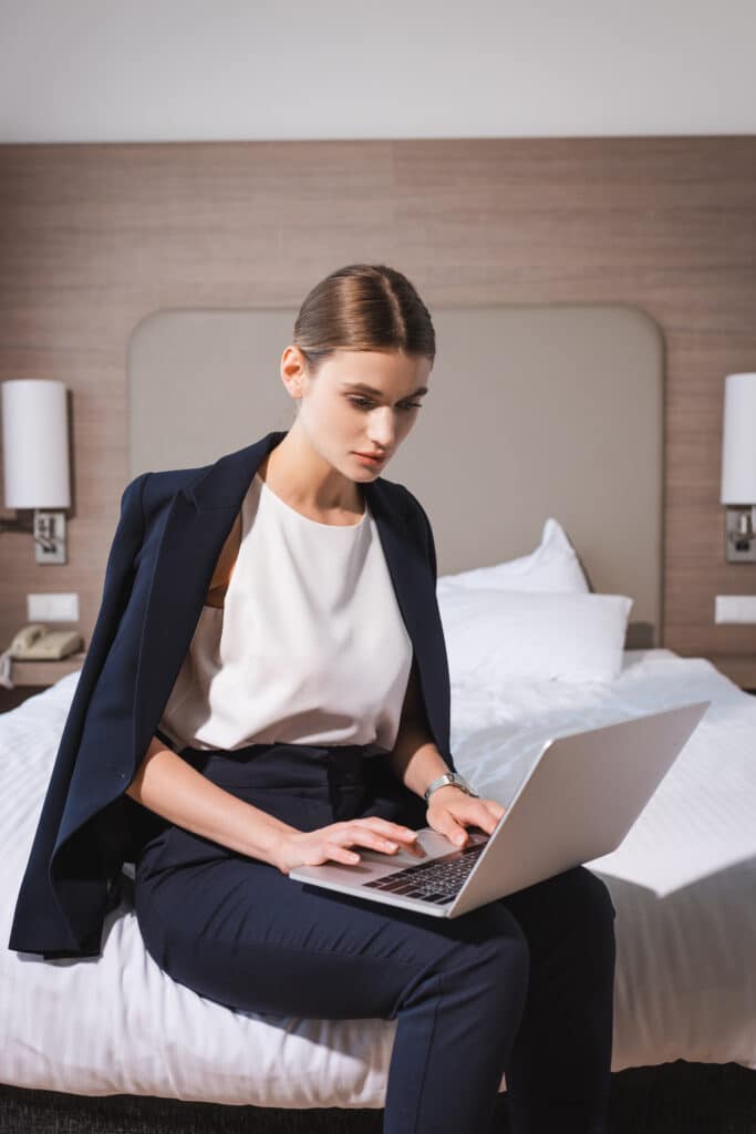Travail : professionnelle utilise son ordinateur portable à l'hôtel. Femme d'affaires assise sur un lit d'hôtel, en blazer bleu marine, travaillant sérieusement sur son ordinateur portable.