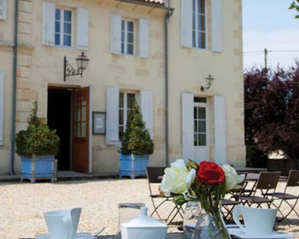 Déjeuner d'été sur une terrasse provençale. Table dressée, roses rouges et blanches, devant un mas traditionnel aux volets clairs.