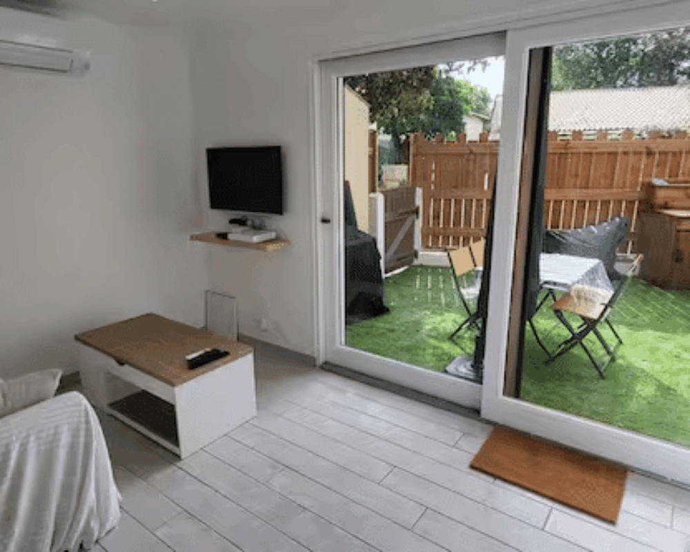 Salon moderne blanc avec canapé, table basse en bois et baie vitrée ouvrant sur un petit jardin avec gazon synthétique.