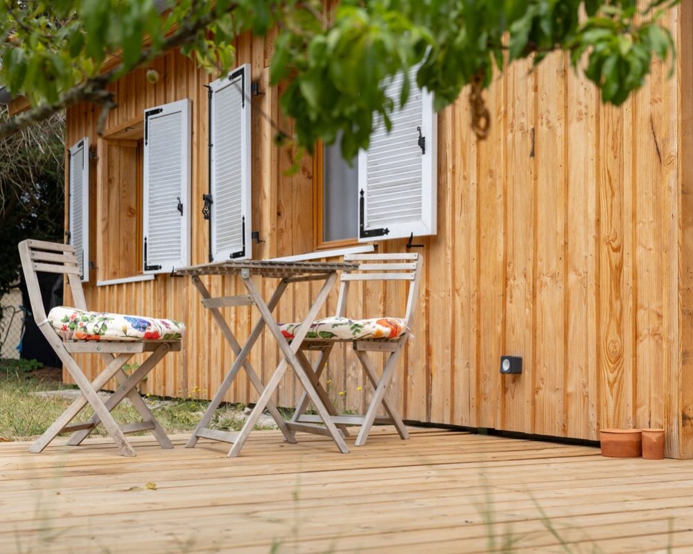 Terrasse d'un chalet en bois avec table, chaises pliantes et volets blancs. Ambiance rustique et paisible.