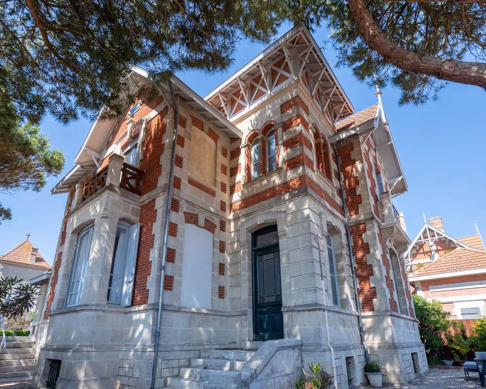 Villa historique en briques et pierre, architecture élégante sous les pins et ciel bleu.